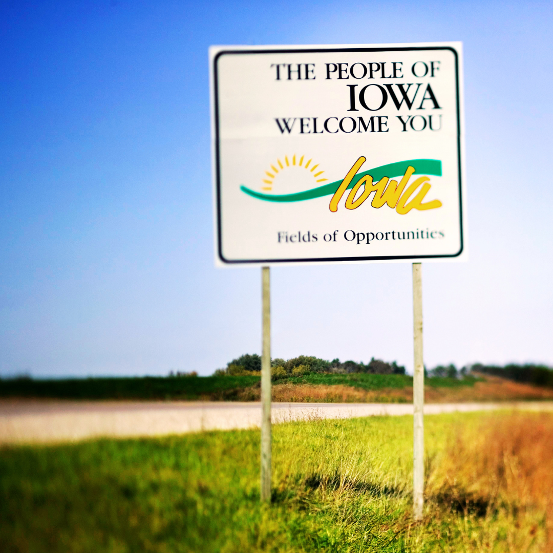 a "welcome to iowa" highway sign set against a backdrop of green vegetation and a blue sky