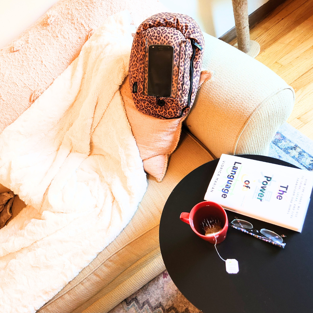 a sunlit couch with a leopard print phone spud resting on the couch. a book titled 'The Power of Language', a red mug of tea, and glasses rest on a small table.